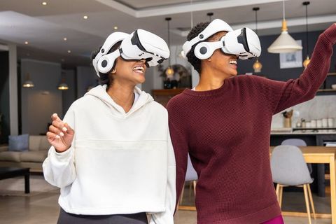 Laughing women wearing vr headsets and pointing while exploring virtual reality at home