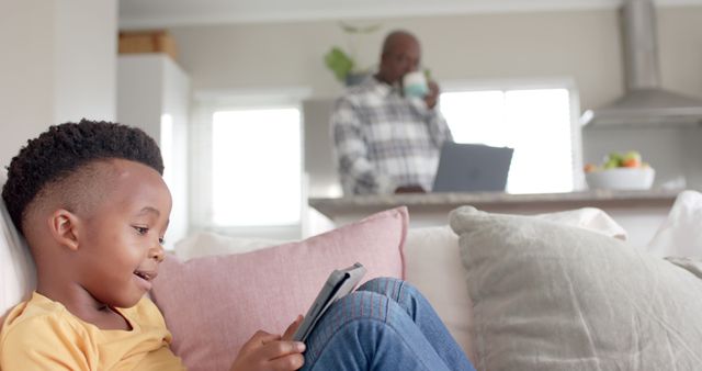 Young boy sitting on a couch enjoying a digital tablet while his parent is working in the background, creating a blend of leisure and work-from-home life. Ideal for use in articles or promotions related to family dynamics, technology, modern parenting strategies, and balanced home lifestyles.