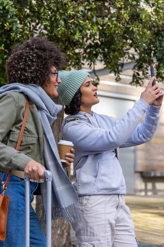 Two friends taking selfie while traveling with luggage and coffee on urban sidewalk