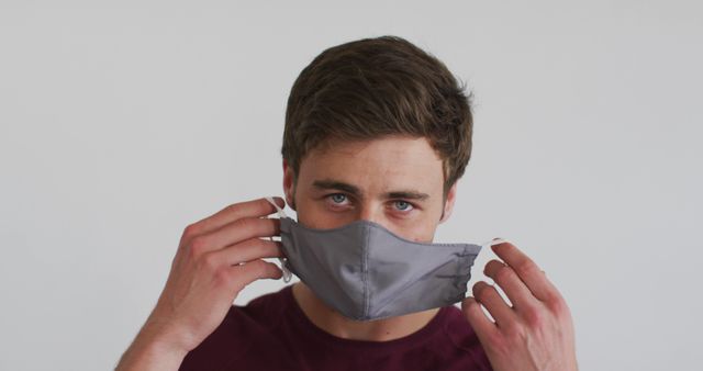 This image captures a young man in a close-up view as he adjusts his face mask against a plain background. Ideal for promoting health and safety measures, pandemic-themed content, or educational materials on proper mask usage.