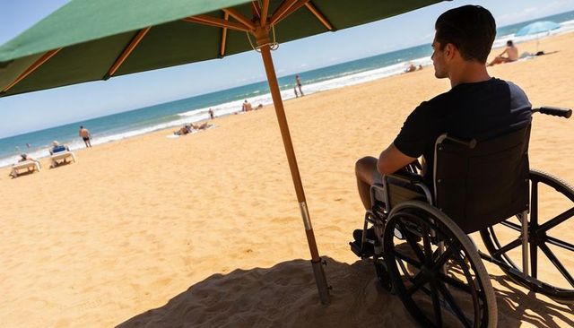 Wheelchair user enjoying ocean view under umbrella on sunny beach, accessible travel