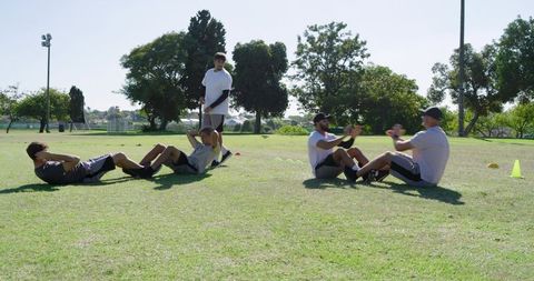 Men training outdoors doing partner sit-ups on grass with coach guiding fitness drill