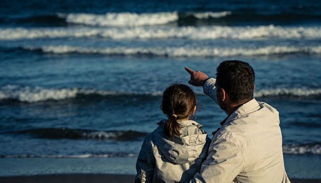 Father pointing to horizon while sitting with daughter on windswept shoreline