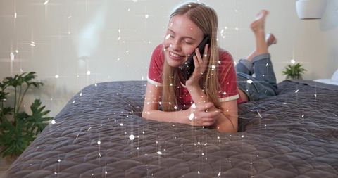 Young woman lying on bed smiling while talking on smartphone in cozy bedroom