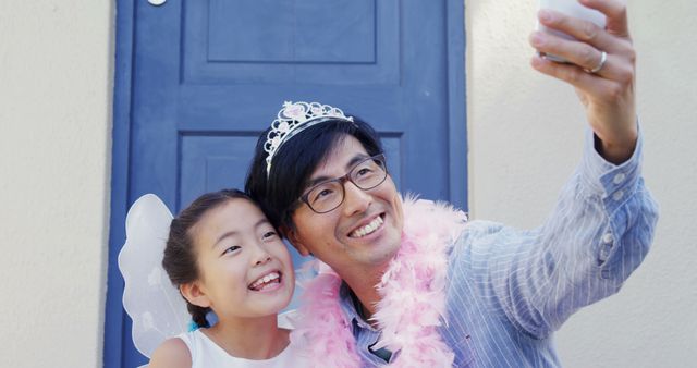 An Asian man and a young girl are capturing a joyful moment with a selfie, both smiling brightly. Dressed in playful attire, the girl as a fairy and the man with a pink feather boa, they share a special bond.