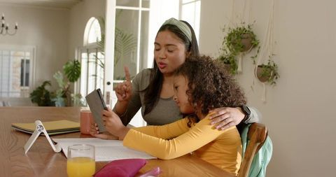 Parent helping child with tablet homework at sunlit dining table with indoor plants