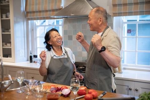 Joyful senior asian couple cooking together at kitchen island wearing matching aprons