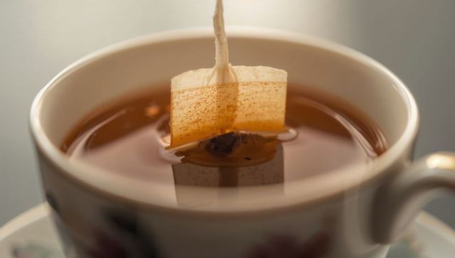 Steeping tea bag releasing amber infusion in porcelain teacup macro close-up, warm beverage