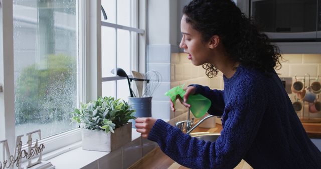 Woman watering a small indoor plant placed on a windowsill in a modern, bright kitchen. She is holding a green spray bottle and dressed in a blue casual sweater. This image perfect for home living articles, houseplant care tips, modern home decor ideas, and lifestyle blogs.