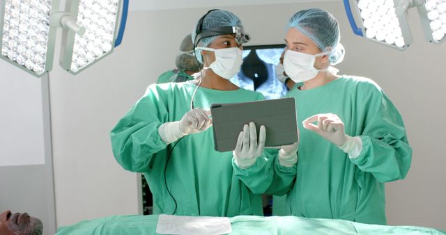 Surgeons in operating room wearing green scrubs, surgical masks, and hair nets discussing medical procedure using digital tablet. Useful for topics related to advanced medical technology, healthcare teamwork, surgery, and modern medical practices.