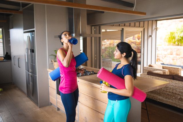 Mother and daughter hydrating after a home workout, holding yoga mats and water bottles in a modern kitchen. Ideal for content related to family fitness, healthy living, home workouts, and active lifestyles.