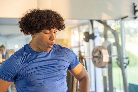 Young man catching breath after heavy squat in bright gym with mirror and loaded barbell