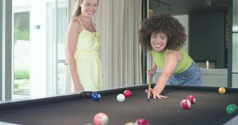 Young african american woman aiming cue while friend watches on modern home pool table