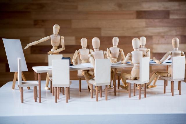 Wooden figurines are seated around a table in a conference room, engaging in a business meeting. One figurine is standing and pointing at a blank presentation board, suggesting a collaborative and strategic discussion. This image can be used for illustrating concepts of teamwork, corporate meetings, business strategy, and leadership in presentations, articles, and marketing materials.