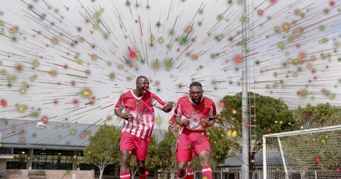 Two soccer players celebrating goal with confetti burst on outdoor grass pitch during victory