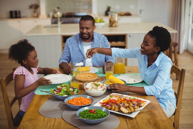 Happy family sitting around a dining table enjoying a meal at home. The scene includes parents and a child, with a variety of healthy foods such as vegetables, salads, and corn on the cob. This image can be used for advertisements or articles promoting family time, healthy eating, and togetherness. Excellent for use in lifestyle blogs, family-oriented websites, and advertisements for food and kitchen products.