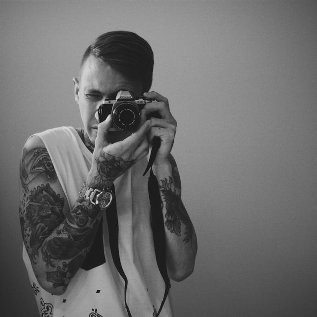 Young man with extensive arm tattoos and wristwatch using vintage camera in a black and white photograph. Artistic pose suggests creativity and passion for photography. Suitable for themes of self-expression, individual's hobbies, creative pursuits, and vintage artistry.