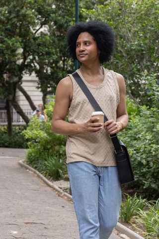 African american man walking with coffee cup and crossbody bag in urban park greenery