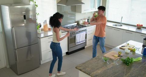 Diverse couple cooking and dancing on granite island in modern sunlit kitchen with avocado prep and