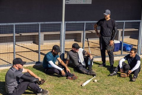 Baseball teammates resting on dugout grass after practice holding gloves and bat in sun