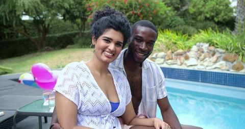 Smiling diverse couple relaxing by backyard pool on sunlit day with float and greenery