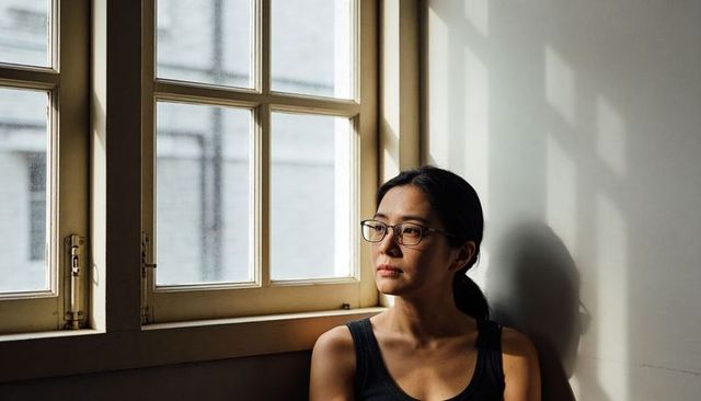 Woman sitting by window, gazing through soft daylight, contemplative minimalist interior portrait