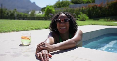 Smiling woman relaxing at pool edge wearing sunglasses and green swimsuit with citrus drink