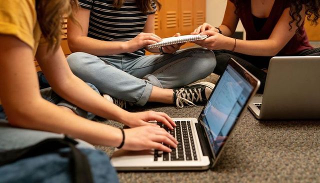Students studying together on hallway floor with laptops and notebooks, collaborating on campus