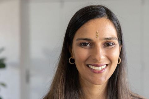 Warm indian woman headshot wearing bindi and gold hoop earrings smiling in bright interior