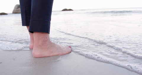Senior Woman's Feet Touching Ocean Waves on Sandy Beach