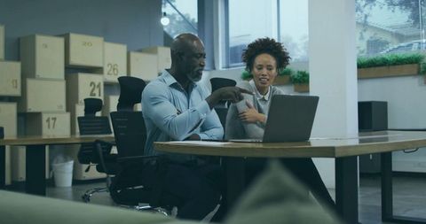 Two colleagues collaborating over laptop at modern office desk surrounded by boxes