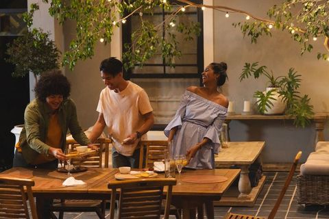 Friends preparing cozy outdoor dinner party on patio at twilight with string lights and plants
