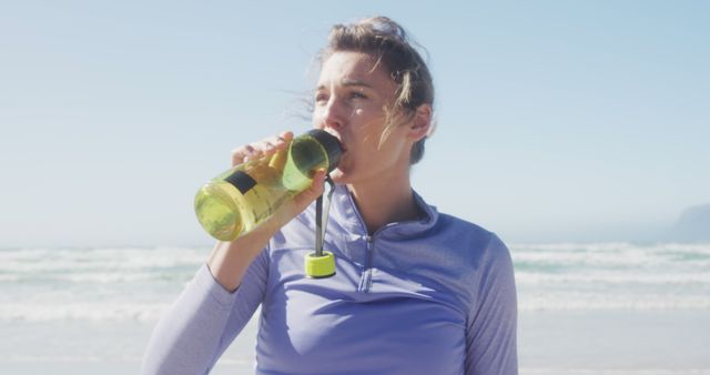 Young woman wearing activewear drinking from water bottle after workout on a sunny beach with ocean waves in the background. Suitable for promoting healthy lifestyle, fitness products, and staying hydrated during outdoor activities.