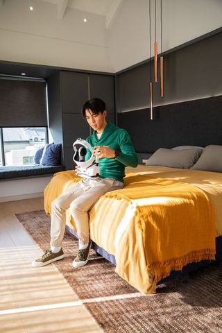 Asian man inspecting vr headset while sitting on bed in modern minimalist warm bedroom