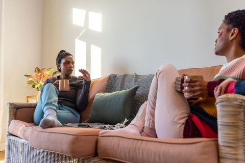 Sunlit conversation between diverse female friends on wicker sofa holding ceramic mugs