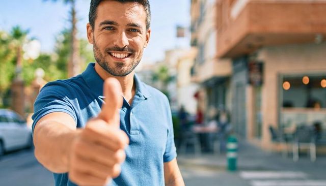 Smiling man giving thumbs-up on sunlit urban street in blue polo showing friendly grin