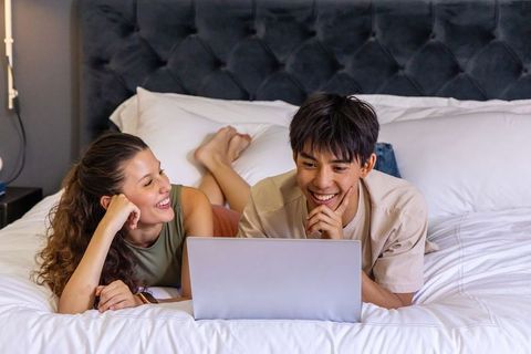 Young couple relaxing on bed using laptop together in cozy modern bedroom for streaming