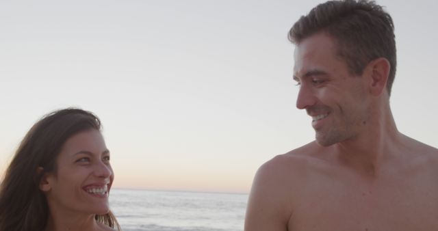 Couple smiling and enjoying each other's company during sunset at beach. Perfect for use in romantic, relationship, and travel related content. Good for ads promoting getaways, vacations, and lifestyle articles focusing on romantic themes.