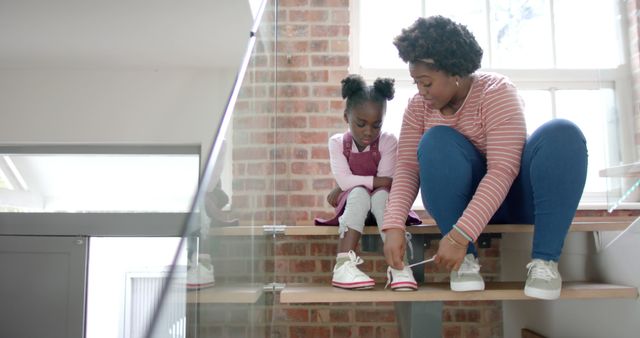 A mother helps her young daughter tie her shoes while they sit on a home staircase. The scene captures a nurturing moment between the parent and child, showing care and tenderness. The home has a modern look with a brick wall in the background. This can be used to illustrate family life, parenting, and the bond between mothers and daughters.