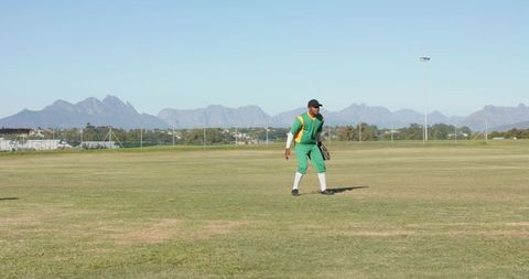 Standing cricketer in green uniform fielding on outfield with mountain backdrop