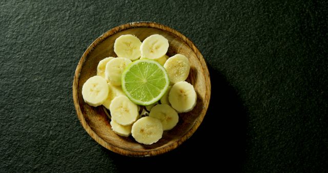 Top view of a wooden bowl filled with banana slices and a lime slice in the center, placed on a dark surface. Perfect for use in blog posts about healthy eating or fruit recipes, advertisements for organic food products, or kitchen decor inspiration.