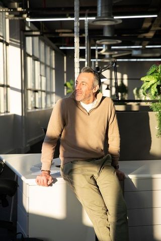 Middle-aged professional sitting on desk in sunlit modern loft office, holding smartphone