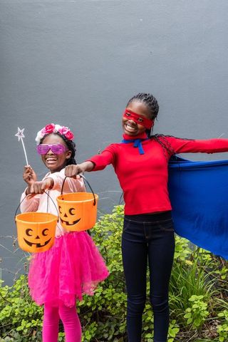 Joyful african american girls wearing superhero cape and pink tutu holding pumpkin buckets