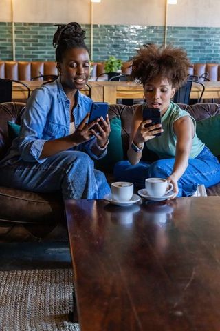 African american friends sharing coffee and scrolling phones on leather sofa in cozy cafe