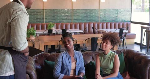African women chatting with server at cozy cafe, ordering coffee, holding phone and tablet