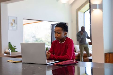 Youth using laptop at kitchen island while African American father walking out on phone