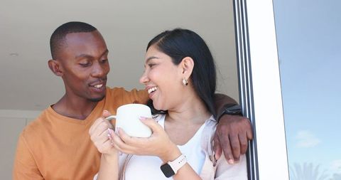 Smiling diverse couple sharing morning coffee by sliding door while showing smartwatch