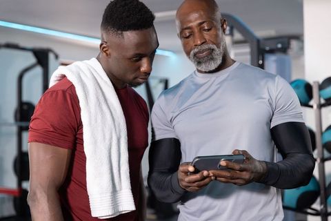 Senior african american coach showing smartphone to male trainee for gym training guidance