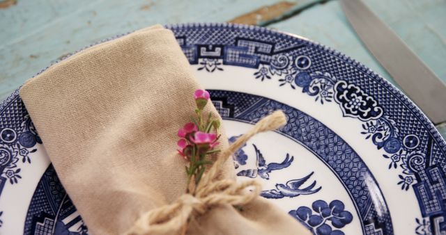 Close-up photograph of an elegant table setting featuring a blue and white ceramic plate, a beige linen napkin decorated with a small bunch of pink flowers, and twine. Perfect for illustrating themes like vintage dining, rustic elegance, decorating ideas, hospitality, festive celebrations, and place settings in culinary contexts.