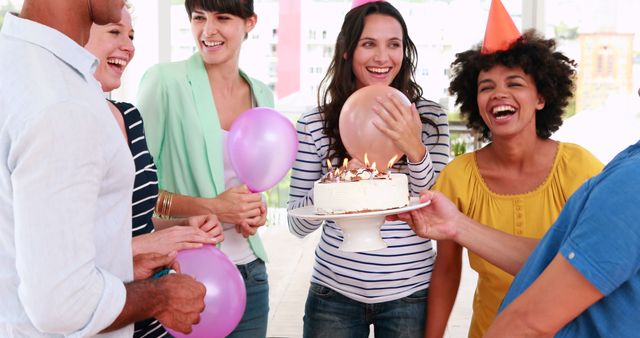 A diverse group of adults celebrates a birthday, with one person holding a cake adorned with lit candles, with copy space. Joy and excitement are evident as balloons add to the festive atmosphere of the gathering.
